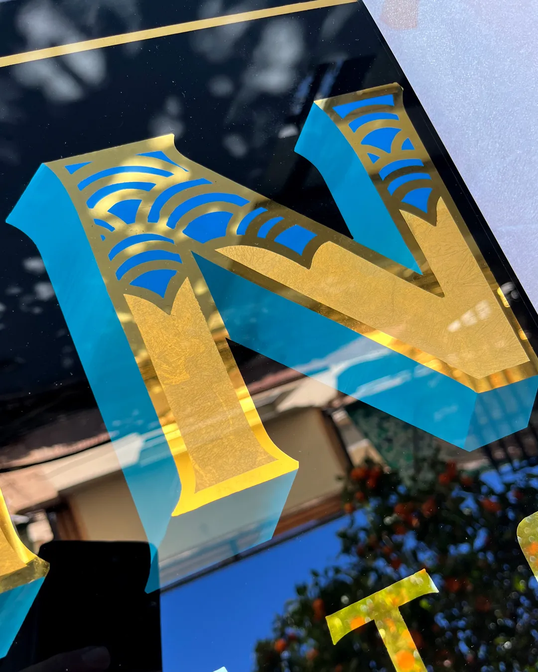 Detalle de letra rotulada a mano en vidrio con pan de oro brillante y mate en rótulo tradicional para estudio de tatuajes en Valencia